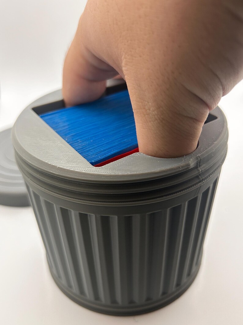May include: A gray, 3D-printed trash can-shaped container with a lid. The container has vertical ridges and holds a stack of blue and red cards. A hand is reaching into the container.