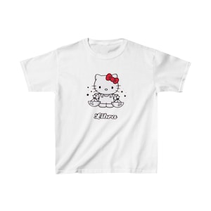 Puede incluir: Camiseta blanca con una imagen de dibujos animados de Hello Kitty vestida como el signo del zodíaco Libra. El texto "Libra" está impreso debajo de la imagen.