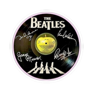 May include: A round sticker featuring a black vinyl record with the band name "The Beatles" in white. The record has the signatures of the band members and the "Abbey Road" album cover. The sticker has a pink border.