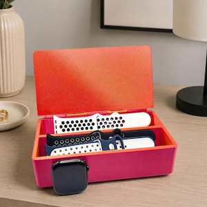 May include: A magenta and orange watch band organizer with several watch bands in white, navy, and black. A black smartwatch is placed next to the organizer. The organizer is on a wooden surface.