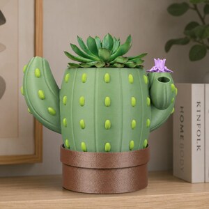 May include: A green cactus-shaped planter with a brown base, holding a green succulent and a small purple flower. The planter has yellow-green accents and is sitting on a wooden shelf. The planter is a decorative item.