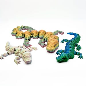 Peut inclure: Trois lézards articulés en jouet de différentes couleurs. Le premier lézard est arc-en-ciel, le deuxième est vert clair et le troisième est brun clair.