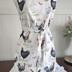 May include: White apron with a chicken and egg pattern. The apron has a black and white chicken, a white chicken, and a brown and white chicken. The apron also has a basket of eggs, a cup of tea, and a sprig of lavender.