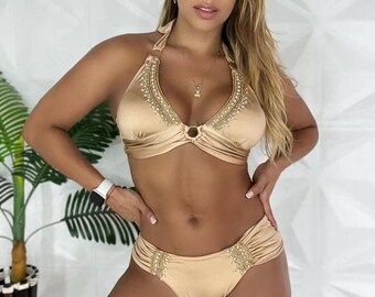 Puede incluir: Un conjunto de bikini dorado. El top de estilo halter y la braguita de cintura baja están adornados con abalorios decorativos. La tela tiene un brillo satinado. El bikini está diseñado para trajes de baño.