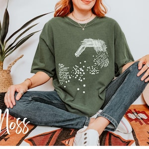 May include: A woman wearing a green oversized t-shirt with a white abstract design. The shirt has the word "Moss" printed on it. She is wearing blue jeans and white sneakers.
