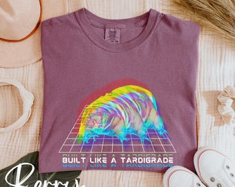Puede incluir: Camiseta morada con un gráfico de un tardígrado en estilo retro con el texto "Built like a tardigrade" impreso en la parte delantera.