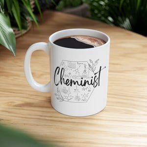 May include: White ceramic coffee mug with a black handle. The mug features a black and white illustration of a hexagon with the word "Chemnist" and chemical formulas and diagrams inside. The mug is filled with black coffee.