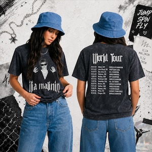 Op de afbeelding: Een antracietgrijs acid-wash T-shirt met een afbeelding van Ilia Malinin met zonnebril op de voorkant en op de achterkant een lijst met World Tour-data en prestaties, gecombineerd met een blauwe spijkerhoed en een blauwe spijkerbroek.
