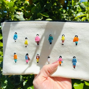 Op de afbeelding: Crèmekleurige canvas etui met een zwarte rits, geborduurd met kleurrijke figuurtjes. De figuurtjes zijn in verschillende kleuren, waaronder blauw, roze, geel en oranje. De etui wordt tegen een groene, bladerrijke achtergrond gehouden.