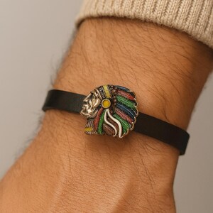 Pode incluir: Pulseira de couro preta com uma cabeça de chefe nativo americano prateada com detalhes de penas coloridas. A pulseira é usada no pulso, mostrando o design detalhado e o artesanato da joia.
