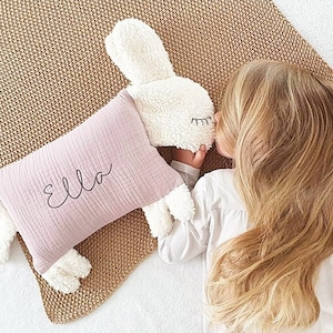 Könnte beinhalten: Ein rosa-weißes Plüsch-Hasen-Kissen mit dem Namen "Ella" bestickt. Das Kissen liegt auf einem braunen gewebten Teppich und ein junges Mädchen hält es.