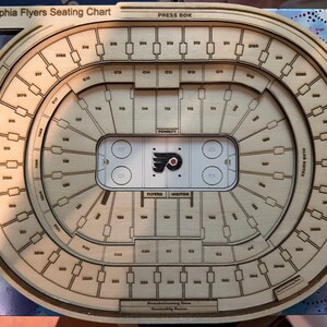 May include: A laser-cut wooden Philadelphia Flyers seating chart featuring an oval arena layout with engraved section numbers, a central hockey rink graphic with the team logo, and labeled areas including the Press Box and Liberty Lofts.
