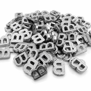 May include: A pile of small, metallic letter "B" charms. The charms are silver-colored and have a textured surface. The letters are scattered across a white background, creating a visually interesting arrangement. These charms could be used for jewelry making or other craft projects.