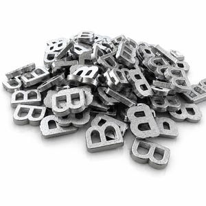 May include: A pile of small, metallic letter "B" charms. The charms are silver-colored and appear to be made of metal. They are scattered on a white surface, with some overlapping. The lighting is bright, highlighting the texture of the metal.