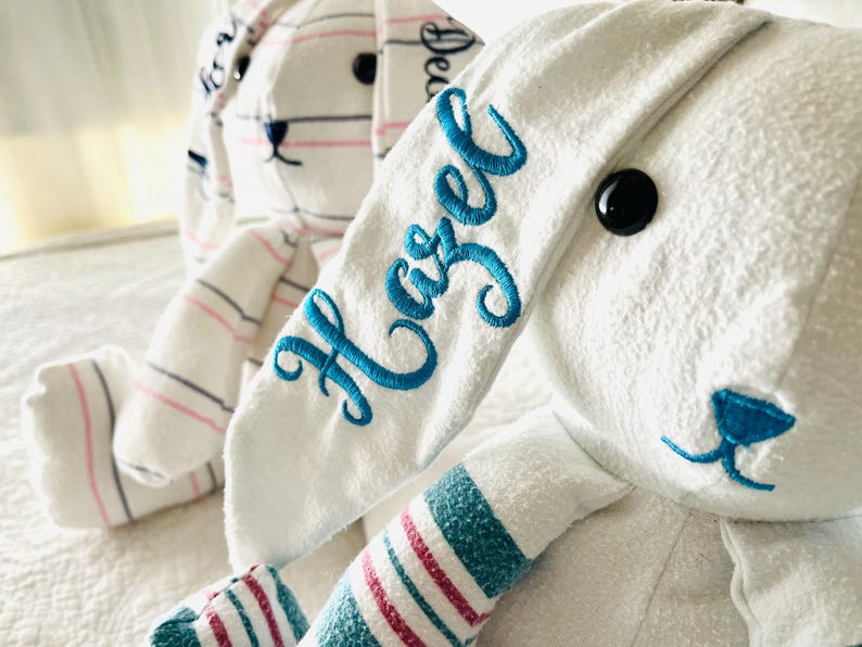 May include: Two plush bunny rabbits. One is white with blue embroidered text that reads "Hazel" on the ear. The other bunny is white with pink and blue stripes. Both have black button eyes and blue accents.