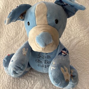 May include: A plush blue dog with white accents, featuring a light blue body and ears with a bunny pattern. The dog has black eyes and a white nose. Text on the dog reads "5-3-18, 6lbs 13oz, 4:41 pm".