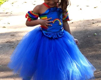 May include: A young girl in a vibrant rainbow-themed costume. The outfit includes a blue satin bodice with a rainbow and star accents, a voluminous blue tulle skirt, and rainbow-striped sleeves. She wears red boots and a blue bow.