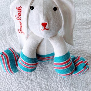 May include: A white plush bunny with long ears, black eyes, and a red nose. The bunny's feet are teal with red and white stripes. The name "Jason Cash" is embroidered in red on one ear. The bunny is a soft toy.