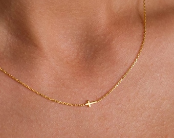 May include: A delicate gold chain necklace featuring a small, simple cross pendant positioned off-center along the thin, polished metal links. The necklace rests against the collarbone, showcasing a minimalist design with a bright, reflective finish.