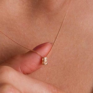 May include: A delicate rose gold-toned chain necklace featuring a small, cylindrical pendant engraved with the capital letter B. The pendant is suspended from a thin, fine-link chain that rests against the collarbone area in a simple design.