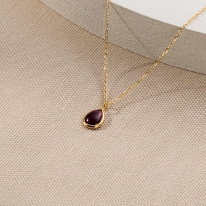 May include: A gold-colored necklace with a teardrop-shaped, dark red pendant. The pendant is set in a gold bezel and hangs from a delicate gold chain. The necklace is displayed on a neutral-toned surface.