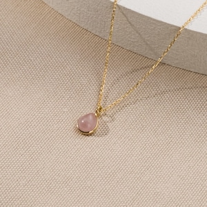 Op de afbeelding: Een delicate gouden ketting met een kleine, gladde, druppelvormige roze edelsteen hanger. De hanger is gezet in een eenvoudige gouden zetting en de dunne, gepolijste gouden ketting is verbonden met de bovenkant van de zetting met een ringetje.