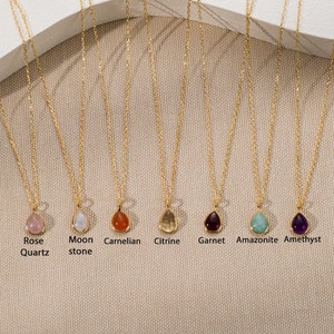 May include: Seven gold necklaces with teardrop-shaped gemstone pendants labeled Rose Quartz, Moonstone, Carnelian, Citrine, Garnet, Amazonite, and Amethyst. Each pendant is set in a gold bezel and hangs from a delicate gold chain, showcasing a variety of natural stone colors.