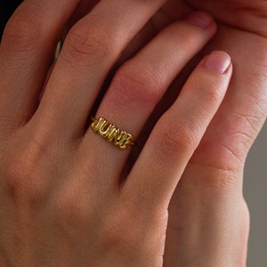 May include: A gold-toned ring featuring the word JUNE in a rounded, three-dimensional, balloon-style font. The letters are connected to a thin, smooth, circular band that encircles the finger, showcasing a polished, reflective metallic finish.