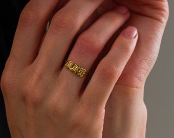 May include: A gold-toned ring featuring the word JUNE in a rounded, three-dimensional, balloon-style font. The letters are connected to a thin, smooth, circular band that encircles the finger, showcasing a polished, reflective metallic finish.