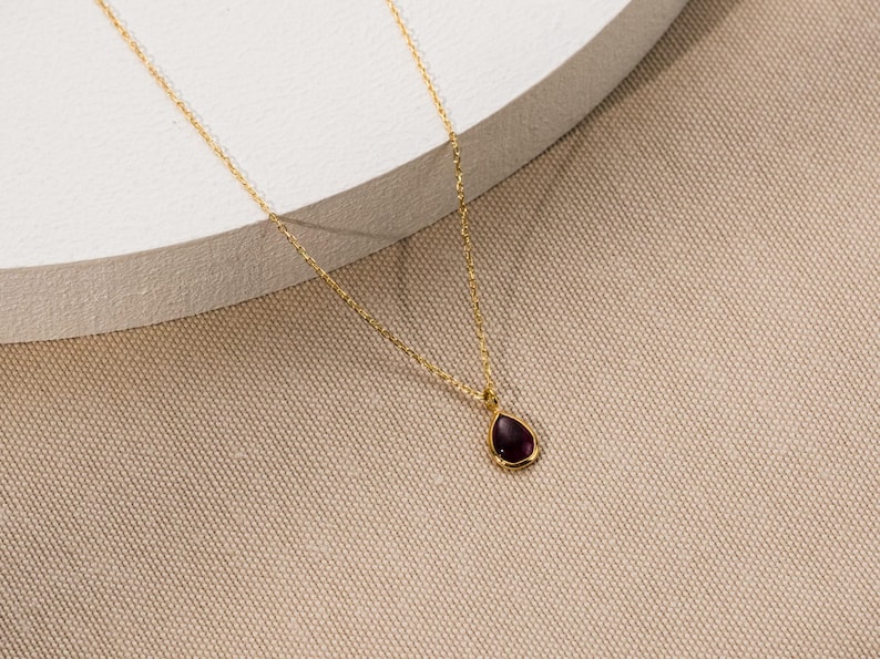 May include: A delicate gold chain necklace featuring a single teardrop-shaped pendant with a deep purple gemstone set in a smooth gold bezel. The fine link chain is crafted from polished gold, providing a simple and elegant look for the jewelry piece.