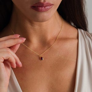 May include: A delicate gold chain necklace featuring a single, small, teardrop-shaped pendant with a deep red gemstone set in a gold bezel. The necklace rests against the collarbone, showcasing a simple, refined design with a polished finish.