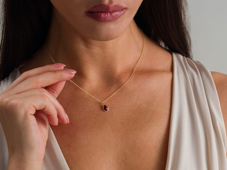 May include: A delicate gold chain necklace featuring a single, small, teardrop-shaped pendant with a deep red gemstone set in a gold bezel. The necklace rests against the collarbone, showcasing a simple, refined design with a polished finish.