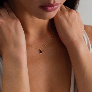 May include: A delicate gold chain necklace featuring a small, teardrop-shaped pendant with a deep purple gemstone set in a gold frame. The necklace rests against the collarbone, showcasing a simple, refined design with a polished metal finish.