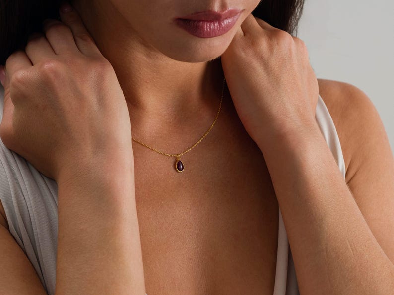 May include: A delicate gold chain necklace featuring a small, teardrop-shaped pendant with a deep purple gemstone set in a gold frame. The necklace rests against the collarbone, showcasing a simple, refined design with a polished metal finish.
