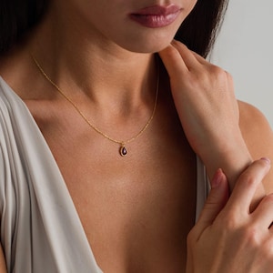 May include: A delicate gold chain necklace featuring a small, teardrop-shaped pendant with a deep red gemstone at its center. The pendant is suspended from a thin, polished gold link chain that rests against the collarbone area of the neck.