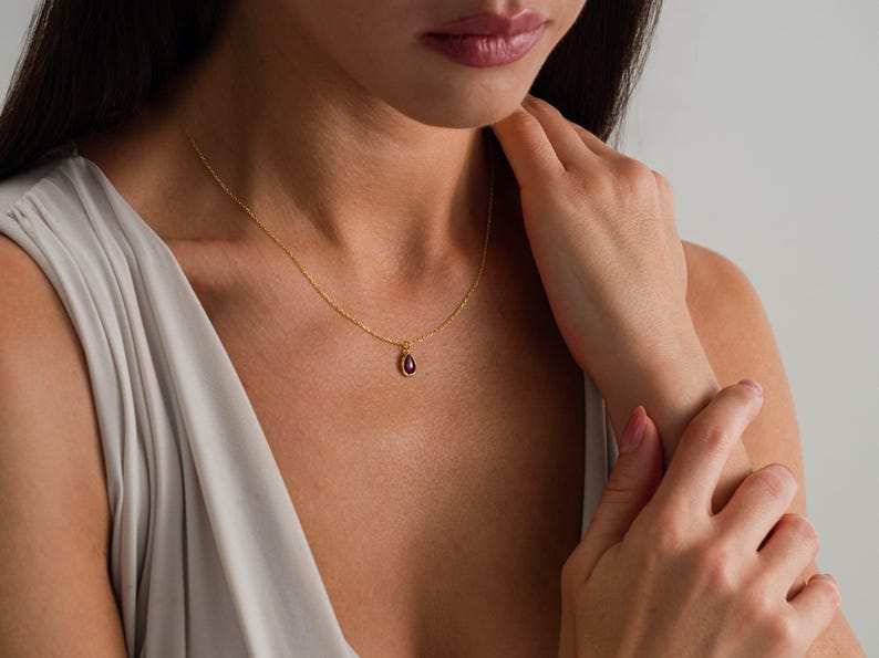 May include: A delicate gold chain necklace featuring a small, teardrop-shaped pendant with a deep red gemstone at its center. The pendant is suspended from a thin, polished gold link chain that rests against the collarbone area of the neck.