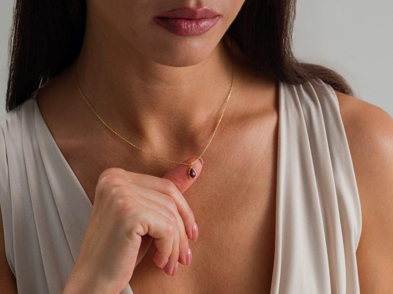 May include: A delicate gold chain necklace featuring a small, teardrop-shaped pendant with a deep red gemstone at its center. The necklace rests against the collarbone, showcasing a thin, polished metal link design and a secure, minimalist setting.