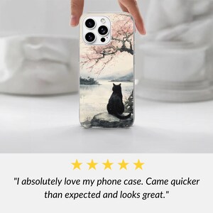 K&ouml;nnte beinhalten: Eine Smartphone-H&uuml;lle mit einer Aquarell-Illustration, die eine schwarze Katze auf einem Felsen an einem See zeigt. &Uuml;ber der Katze erstreckt sich ein Baum mit rosa Kirschbl&uuml;ten. Die H&uuml;lle hat eine Aussparung f&uuml;r ein Dreifach-Kamerasystem.