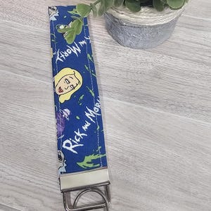 May include: A blue fabric keychain featuring characters and text from the animated series "Rick and Morty." The keychain has a silver metal clasp and ring. The background includes a small potted plant.