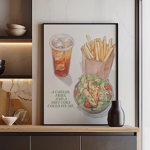 Puede incluir: Una ilustración de acuarela de una ensalada César, patatas fritas y un vaso de té helado. La ilustración está enmarcada y colgada en una pared de una cocina moderna con armarios oscuros y suelo de madera. El texto debajo de la ilustración dice "Una César, patatas fritas y una coca-cola light podrían arreglarme."