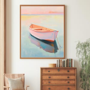 Puede incluir: Una impresión artística enmarcada de un pequeño bote flotando en el agua. El bote está pintado en tonos de rosa y naranja, con un casco blanco. El agua refleja el bote y los colores suaves del cielo. La obra de arte se exhibe en una habitación con muebles de madera.