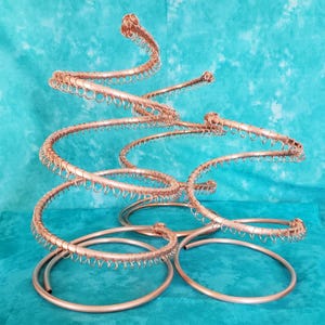 May include: Copper-colored metal sculpture with a spiral design. The sculpture has multiple curved arms and circular bases, creating a dynamic and artistic form. The piece is set against a teal background.