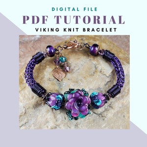 以下が含まれることがあります： 紫とターコイズのバイキング編みブレスレットに花チャームが付いたもの。ブレスレットはワイヤーとビーズで作られています。花チャームはガラス製で、紫とターコイズの色合いです。