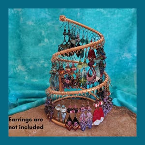 May include: A copper-colored, spiral earring display stand filled with various colorful earrings. The stand is on a brown surface against a teal background. The text "Earrings are not included" is visible at the bottom.
