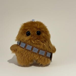 Op de afbeelding: Een bruin, harig pluche speelgoed met grote zwarte ogen en een bandolier. Het speelgoed heeft een lichtblauwe accent op het hoofd en een bruine en grijze band om de torso. Het speelgoed is een Star Wars-personage.