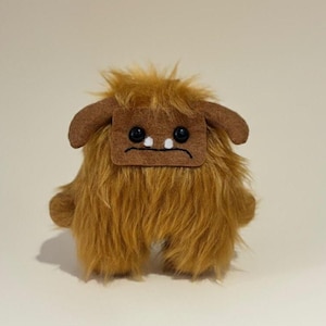 May include: A handmade plush toy in the shape of a furry brown monster. It has a square face with two black button eyes, a stitched frown, and two small white dots. The toy has brown ears and a shaggy body.