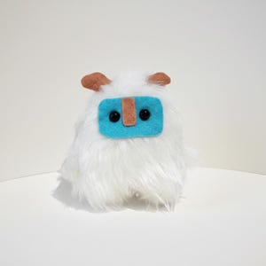 Può includere: Un peluche bianco a forma di mostro con una consistenza pelosa. Presenta un rettangolo turchese con una striscia marrone e due occhi a pois neri. Il giocattolo ha due piccole forme marroni a forma di orecchio sulla parte superiore.