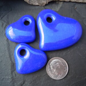 May include: Three blue ceramic heart-shaped charms with holes for hanging. The charms are different sizes and are arranged on a dark gray surface.