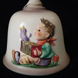 May include: A ceramic bell featuring a 3D image of a boy playing an accordion with a bird perched on his shoulder. The boy is wearing a green shirt, red trousers, and a red bandana. The bell is cream coloured with brown accents.