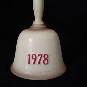 May include: A white ceramic bell with a brown rim and a red inscription of "1978". The bell has a white handle and a signature that reads "M.I. Hummel".